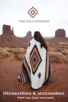 a woman wrapped in a blanket sitting on top of a red dirt field next to mountains