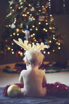 a baby sitting in front of a christmas tree wearing a reindeer antlers headband