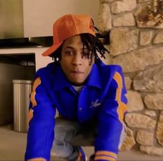 a young man with dreadlocks wearing a blue jacket and orange hat sitting in front of a stone wall