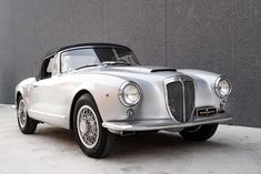 an old silver sports car parked in front of a gray wall with the hood down