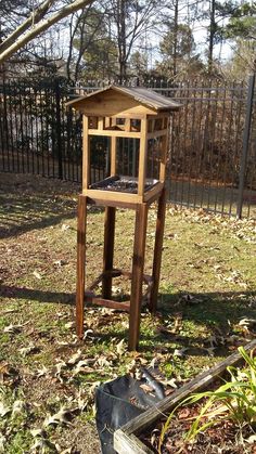 a wooden bird feeder in the middle of a yard next to a fence and tree