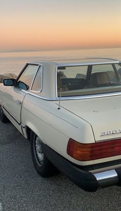 an old white car parked on the side of the road next to the ocean at sunset