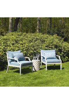 two chairs and a table sitting in the grass next to some bushes with trees behind them