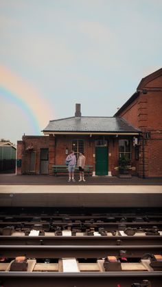 two people standing in front of a train station with a rainbow in the sky behind them