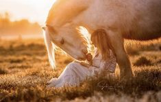 a woman is sitting in the grass next to a white horse with its head down