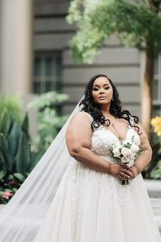 a woman standing in front of a building wearing a wedding dress and holding a bouquet