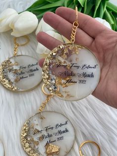 two white and gold necklaces in the palm of a person's hand with flowers behind them