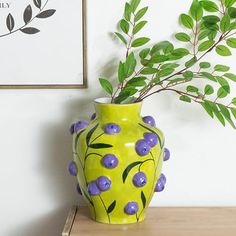 a yellow vase with blue berries on it sitting on a table next to a plant