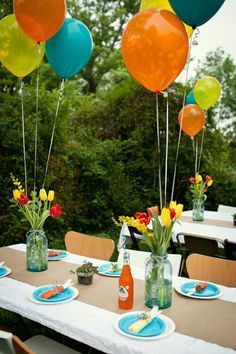 an outdoor table set up for a party with balloons in the air and plates on the ground