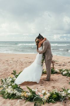 a bride and groom kissing on the beach in front of an arch of greenery