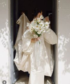 a woman in a white dress is holding a bouquet of flowers and looking out the door