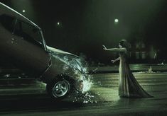 a woman in a long dress standing next to a car on a wet road at night