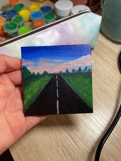 a hand holding up a small painting of a road in the middle of a field