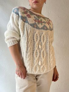 a woman standing in front of a white wall wearing a sweater with an intricate design