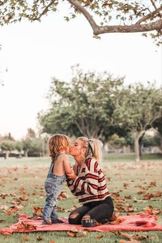 a mother kissing her child while sitting on a blanket in the grass with leaves all around