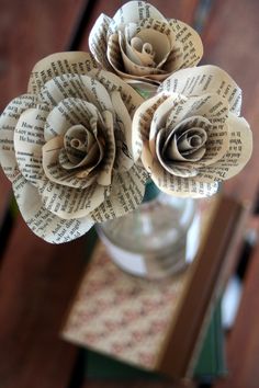 three paper flowers are in a vase on a wooden table with an old book page