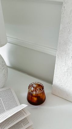 a glass with ice sits on a table next to an open book and a lamp