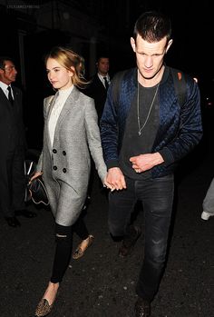 a man and woman walking down the street at night with people in the background holding hands