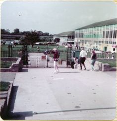 an old photo of people walking around in front of a building with green grass and fence