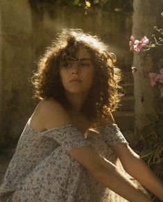 a woman with curly hair sitting on the ground in front of some flowers and bushes