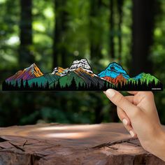 a hand holding up a piece of paper with mountains in the background and trees around it