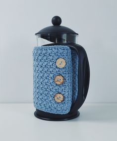 a blue crocheted coffee pot cover with buttons on the front and bottom, sitting next to a black teapot holder