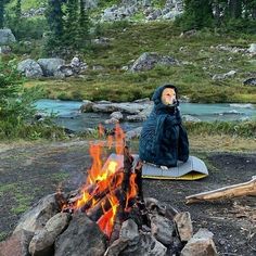 a man sitting next to a fire in the middle of a field with rocks and trees