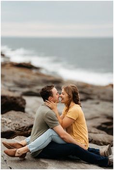 a man and woman sitting on rocks near the ocean kissing each other with their noses touching