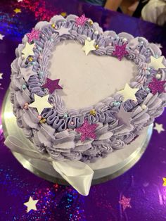 a heart shaped cake with purple icing and stars on the top is sitting on a table