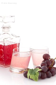 two glasses filled with liquid sitting next to some grapes and a bottle of wine on a white surface