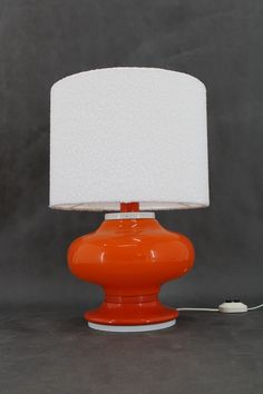 an orange table lamp with a white shade on the base and a cord plugged in