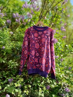 a purple and red sweater hanging on a tree branch in front of some bushes with purple flowers