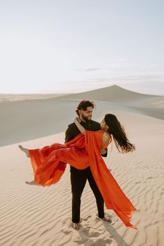 a man and woman dancing in the sand with an orange scarf around their necks, on top of a dune