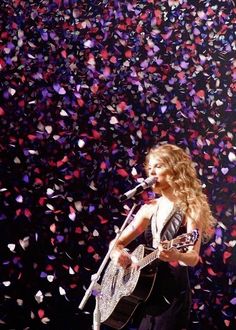 a woman singing into a microphone and holding a guitar in front of confetti