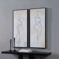 two framed art pieces on a wall above a table with a book and candle holder