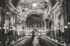 a bride and groom standing in the middle of a large hall with tables set up for dinner