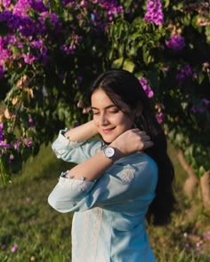 a woman standing in front of purple flowers with her arms around her neck and looking at the camera