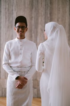 a man and woman standing next to each other in front of a stone wall wearing white clothing