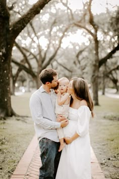 a man and woman are holding their baby while standing on a brick path in front of trees