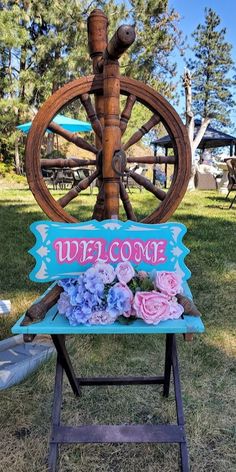 a sign that says welcome with flowers in front of a wheel on the grass and picnic tables behind it