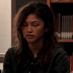 a woman with long hair sitting in front of a book shelf and looking at the camera