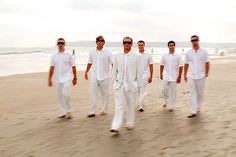 a group of men standing on top of a sandy beach next to the ocean in white outfits