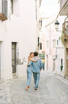 a man and woman walking down an alley way in the city hugging each other with their arms around each other