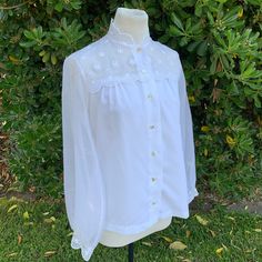 This is a super pretty sheer with blouse with floral embroidery  It buttons up with pearly white buttons Has a bit of a mock neck  The detailing is absolutely beautiful The tag is so worn that the size and material are no longer legible. I can't make out the tag at all. It would probably best fit a size small but please see measurements  It's in great condition, the only imperfections I noticed were in the sleeves, maybe a tiny snag on each sleeve (see pics). Measurements as follows: Pit to pit Classic White Blouse With Pearl Buttons, White Blouse With Floral Embroidery And Peter Pan Collar, White Button-up Blouse With Pearl Buttons, Feminine Button-up Blouse With Floral Embroidery, White Sheer Button-up Blouse, Vintage Sheer Blouse For Spring, Elegant Embroidered Blouse With Peter Pan Collar, Classic Blouse With Floral Embroidery For Spring, Spring Wedding Button-up Blouse