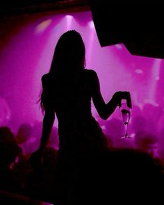 the silhouette of a woman holding a wine glass in front of a stage with purple lights