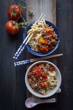 two bowls filled with noodles and meat on top of a wooden table next to tomatoes