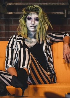 a woman with makeup and hair sitting on a couch wearing striped clothes, holding her hands behind her back