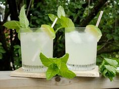 two glasses filled with ice and mint sitting on top of a wooden table next to trees