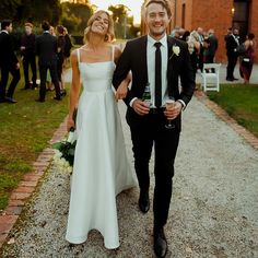 a man and woman walking down a path holding wine glasses in their hands as the sun sets behind them