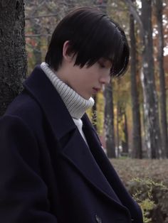 a man standing next to a tree wearing a coat and turtle - neck sweater in the woods